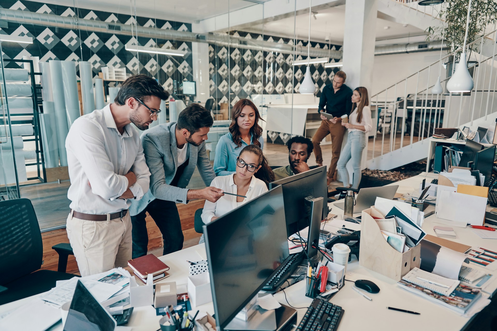 A diverse team of colleagues gathered around a monitor in a busy open office, collaborating